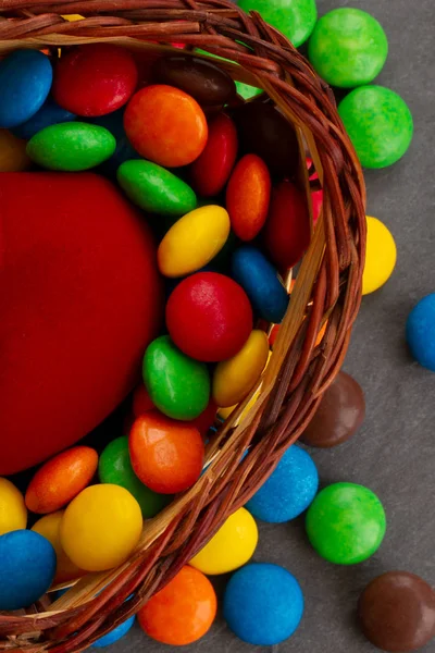 Portrait photo of a basket with sweets. Candy bowl with lots of multicolored sweets and treats. A half of red velvet heart among a set of bright glazed dragees.