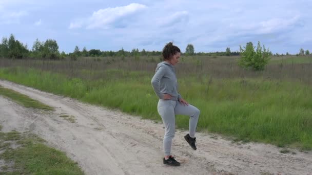 En ung vacker sport Kvinna knådar sina vrister, knän och bäckenleder i den friska luften mitt på en grusväg utanför staden. Långsam panorama. — Stockvideo