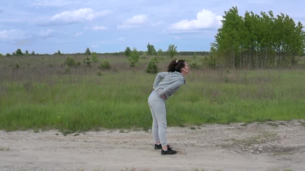 Uma jovem bonita esportes Feminino faz movimentos rotacionais na parte inferior das costas e agachamentos no ar fresco no meio de uma estrada de terra fora da cidade. Panorama lento . — Vídeo de Stock