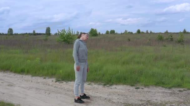 A young pretty sports Female kneads her neck and shoulders in the fresh air in the middle of a dirt road outside the city. Slow-mo panorama. — Stock Video