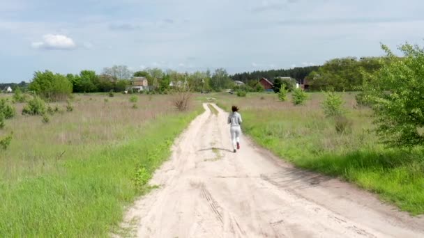 Gri eşofmanlı genç bir kadın çayırların arasında tozlu toprak bir yolda koşuyor. Yavaş Hareket. — Stok video