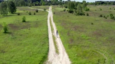 Gri eşofmanlı genç bir kadın çayırların arasında tozlu toprak bir yolda koşuyor. Ağır çekim. Üst Hava Aracı Görüntüsü.