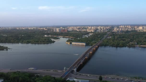 Luftaufnahme eines breiten Flusses mit einer Brücke vor dem Hintergrund von Häusern einer entfernten Stadt Kiew, Ukraine. Abendliches Stadtpanorama. — Stockvideo