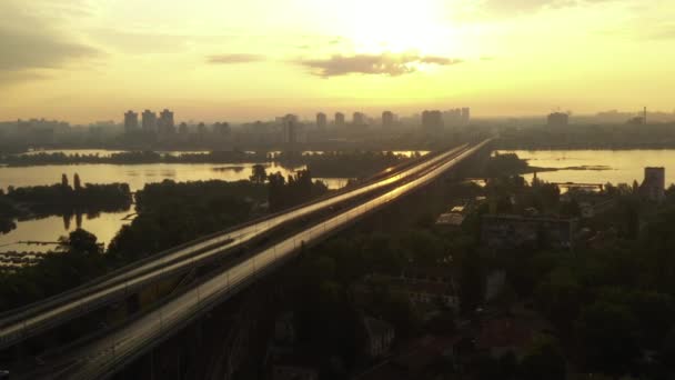 Drona filmowa kręcona w pobliżu dużego długiego mostu z rzadkimi samochodami o świcie w złotej godzinie. Drone lot w pobliżu mostu nad rzeką z wyspą w promieniach wschodzącego słońca. — Wideo stockowe