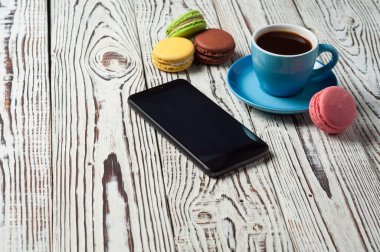 Cep telefonu yakınında siyah kahve Tam mavi fincan ve eski rustik yıpranmış beyaz ahşap masa üzerinde dağınık renkli bütün macaroons
