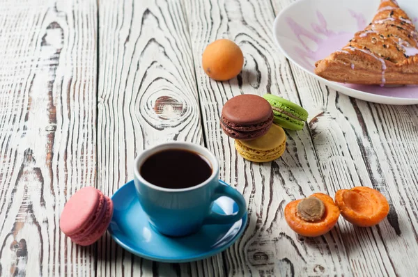 Full ceramic cup of hot black coffee near baked sweet bun in form of triangle with purple cream, apricot, macaroons lies on white weathered wooden table