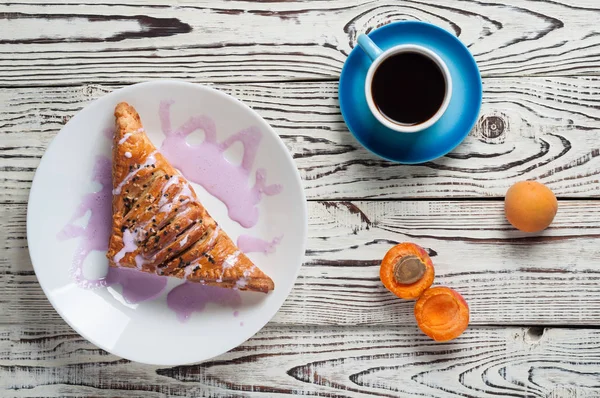 Full ceramic cup of hot black coffee near baked sweet bun in form of triangle with purple cream and apricot lies on white weathered wooden table