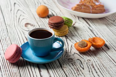 Mor krem, kayısı, macaroons ile üçgen şeklinde pişmiş tatlı topuz yakın sıcak siyah kahve tam seramik fincan beyaz yıpranmış ahşap masa üzerinde yatıyor