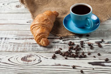 Çuval bezi ve eski beyaz rustik ahşap masa üzerinde taze siyah kahve tam mavi seramik fincan yakın bir bütün pişmiş kruvasan