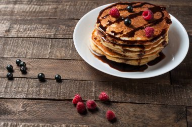 Çikolata krema, ahududu, frenk üzümü ile ev yapımı krep yığını yuvarlak beyaz seramik plaka ve eski rustik ahşap masa yatıyor. Metin için alan kopyalama