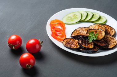 Beyaz seramik plaka ızgara patlıcan süslemeli salatalık, yaprak maydanoz, biber dilimleri dolu çizik koyu beton üzerinde bütün domates yakın yatıyor