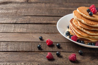 Taze ahududu ile ev yapımı krep yığını, frenk üzümü yuvarlak beyaz seramik plaka ve eski rustik ahşap masa yatıyor. Metin için alan kopyalama