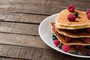 Taze ahududu ile ev yapımı krep yığını, frenk üzümü yuvarlak beyaz seramik plaka ve eski rustik ahşap masa yatıyor. Metin için alan kopyalama