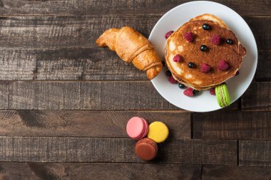 Taze ahududu, kruvasan yakınında frenk üzümü ile ev yapımı krep yığını, macaroons yuvarlak beyaz seramik plaka ve eski rustik ahşap masa yatıyor. Metin için alan kopyalayın. Üstte görüntü