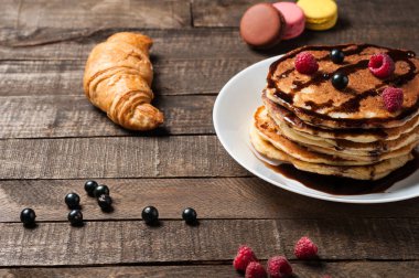 Çikolata krema, ahududu, frenk üzümü, kruvasan, macaroons ile ev yapımı krep yığını yuvarlak beyaz seramik plaka ve eski rustik ahşap masa yatıyor