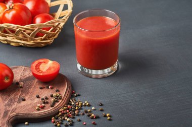 Dolu bir bardak taze meyve suyu olgun domateslerle dolu tahta sepet ve koyu betonun üzerinde duran biber baharatlarıyla dolu tahta kesme tahtası. Metin için boşluk