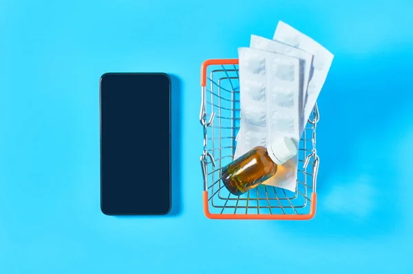 Smartphone near different medicaments in market basket on blue background. E-health, e-medicine, payment for services concept. Online purchasing