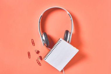 Kablolu, boş kağıt defter ve turuncu arka planda diğer kırtasiye malzemeleriyle modern beyaz kulaklıklar. İş ya da okul konsepti. Uzayı kopyala Üst görünüm
