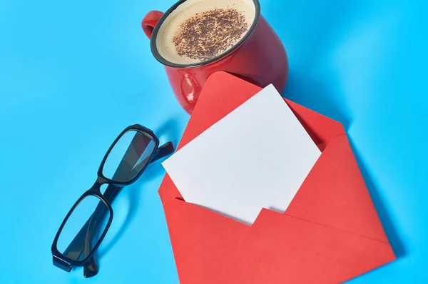 Red envelope with empty paper sheet, full cup of coffee and glasses lies on blue desk. Close-up