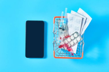 Smartphone, mavi arka plandaki market sepetindeki farklı ilaçların yanında. E-sağlık, e-tıp, hizmet için ödeme konsepti. Çevrimiçi satın alma