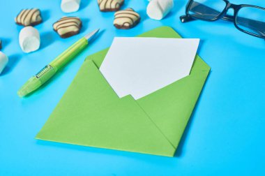 Çizgili şekerler, yeşil zarfın yanında şekerleme, boş kağıt çarşaf, gözlük, mavi masanın üstünde kalem. Aşk mektubu konsepti. Yakın plan.