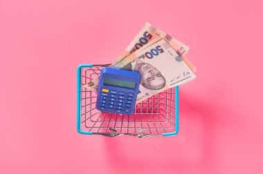 Ukrayna Hryvnia banknotu ve mavi hesap makinesi. Pembe arka planda metal market sepeti. Satın al ya da sat. Döviz kurları kavramı