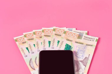 Pembe arka planda akıllı telefon ve Ukrayna parası. Mobil bankacılık kavramı. Evden online alışveriş. Uzaklık denetimi mali denge