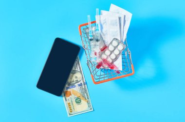 Market sepetindeki farklı ilaçların yanında akıllı telefon ve mavi arka planda 100 dolarlık banknot. E-sağlık, e-tıp, hizmet için ödeme konsepti. Çevrimiçi satın alma