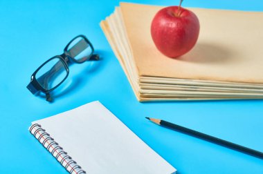 Bir yığın boş dergi, gazete ya da bazı belgeler ve kırmızı elma, not defteri, kalem, mavi masadaki gözlükler. İş, eğitim ya da okula dönüş kavramı. Gazeteci ya da sekreterin işyeri