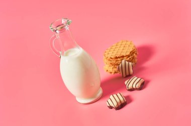 Bir sürahi taze süt, yoğurt ya da ince waffle 'ların yanında kefir ve pembe arka planda çikolata şekeri.