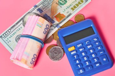 100 dolarlık banknot ve hesap makinesinin yanındaki Ukrayna parası pembe arka planda.