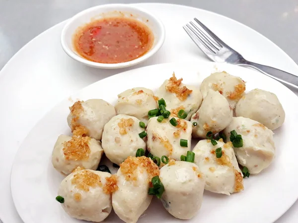 boiled fish balls in a white plate with a small bowl of spicy fish ball sauce, decorated with some vegetable and deep fried garlic on top