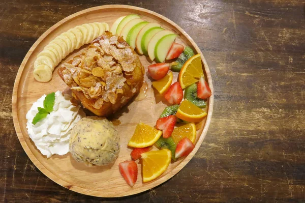 Tatlım meyveli tost ekmeği bir tabak dondurma bir kepçe ile süsleyin, kamçı krema, muz, portakal, çilek, elma, kivi meyve ve Mısır pul. Kahverengi ahşap masanın üzerinde               