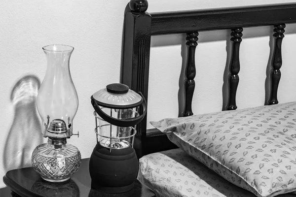 Vintage flat-wick kerosene oil lamp and modern battery operated lamp on bedside table, bed headboard, pillow, mattress, bedroom.