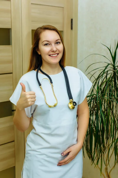 Medical worker with stethoscope smiling, showing thumb up and looking at camera. Cheerful doc gesturing like. Young female nurse with stethocope shows thumb up. Attractive doctor woman show thumbs up
