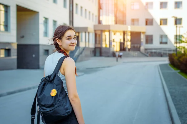 Rear view beautiful brunette with a black backpack behind going to College. A young pretty student looks back at the camera. A beautiful girl going to school. Concept of the Day of knowledge.