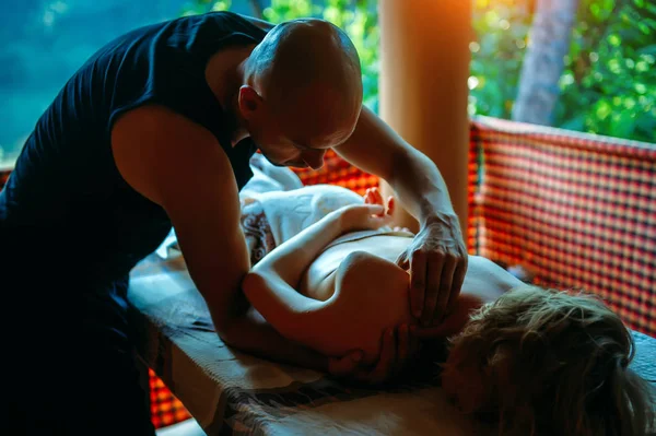 Non-traditional wellness massage outdoor. Young slender masseur massages a woman's shoulder, hand and shoulder blade. Male make therapeutic massage for girl on a massage couch.