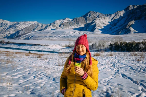 Beautiful girl in red hat holds glass on the background of snow-capped mountains on frosty sunny day. Cheerful young woman enjoying Christmas vacation in the mountains, looking at camera and smiling.