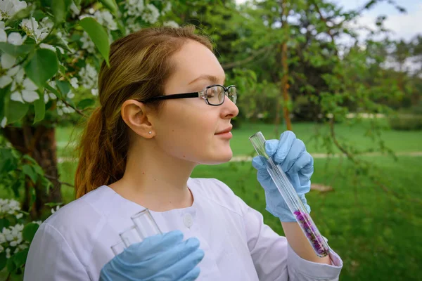 Beautiful female scientist in medical gloves inhales aroma from test tube with flower petals on the background of blooming tree in botanical garden, close-up. Creation of a perfume, natural cosmetics.