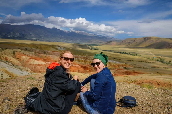 Two young girls in sunglasses sitting on hill look at camera and smile. Women travelers are photographed against the beautiful mountains on sunny day. Active recreation and adventure.