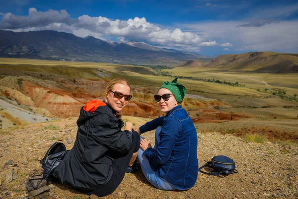 Two young girls in sunglasses sitting on hill look at camera and smile. Women travelers are photographed against the beautiful mountains on sunny day. Active recreation and adventure.