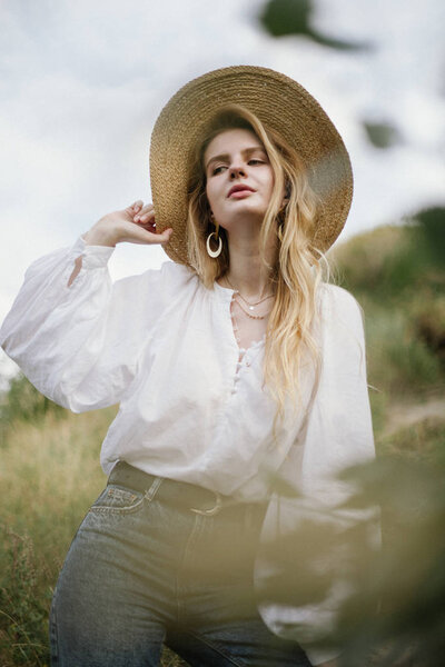 stylish woman in straw hat and white linen blouse and jeans posing on meadow at countryside