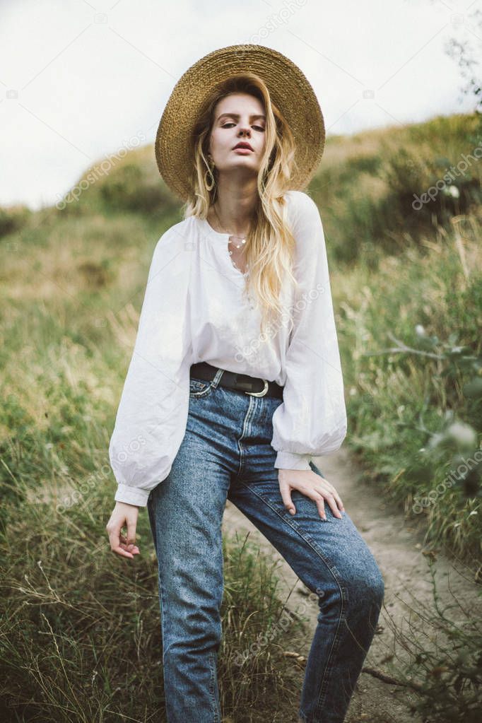 stylish woman in straw hat and white linen blouse and jeans posing on meadow at countryside