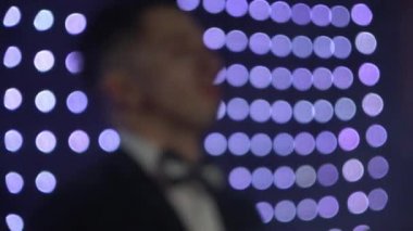 Siyah kravatlı genç bir adam, flaşlı ampullerin arka planına karşı bir sentezleyiciyle oynuyor.