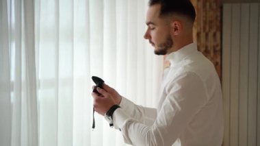 Sakallı bir adam beyaz gömlekli pencerenin yanında duruyor ve papyonu inceliyor.