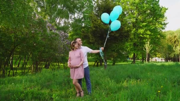 En man med en gravid kvinna som står i parken och håller blå ballonger på våren — Stockvideo