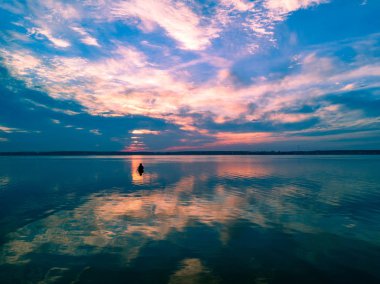 Gölde renkli doğal gün batımı havadan drone panoramik görünümü, suda güneş yansıması, rüzgar olmadan yağmurdan sonra bulutlu gökyüzü, merkezinde yalnız balıkçı, Rusya