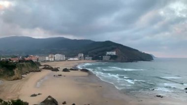 Ispanya 'nın Northen sahilinin Timelapse; sörf spot ve dalgalar ile güzel Atlantik Okyanusu Plajı; kayalık kayalıklar ve bir arka planda nountains; yağmur bulutlar ve güneş ile akşam gökyüzü; kış tatil şehri