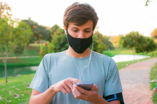 Spor giyimli, elinde telefon kulaklığı ve parkta maskesi olan genç bir adam..