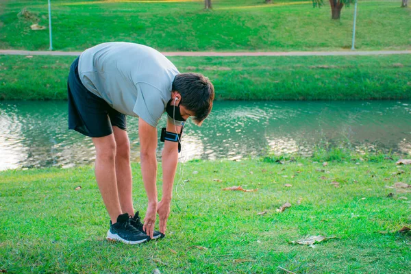 Spor kıyafetleri ve maske takmaya başlamadan önce yeşil bir parkta esneme hareketleri yapan genç bir adam..